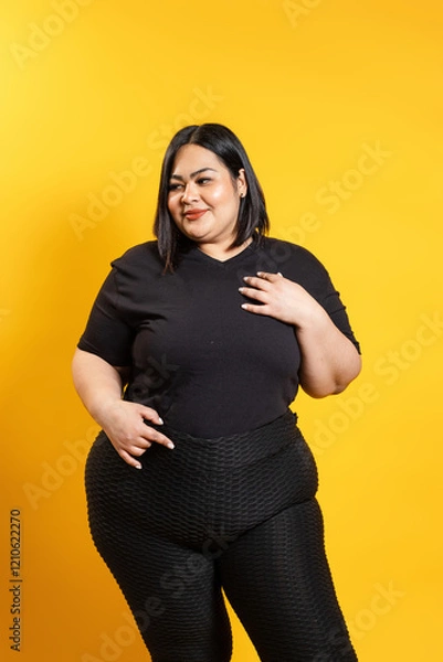 Fototapeta Plus size model posing with hand on chest, embracing body positivity on yellow background