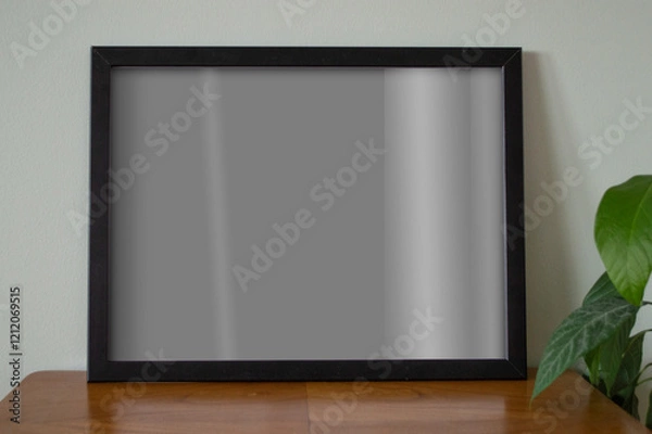 Fototapeta Mockup visualization. Black empty frame standing on wooden table alongside green plant. Poster mock up. Wall art