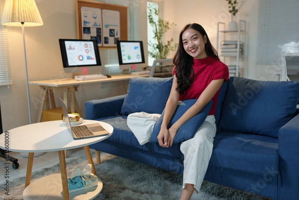 Fototapeta Young asian businesswoman relaxing on the sofa in her living room after a long day of working from home, enjoying the comfort and peace of her home office setup