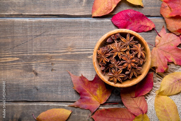 Fototapeta Star anise in a wooden bowl on a wooden table with colorful autumn leaves around. Top view with copy space.