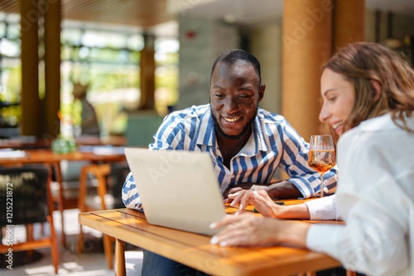 Fototapeta A young Black male and Caucasian female entrepreneur couple engage in work on a laptop at a tropical bar. The setting includes warm sunlight and relaxed ambiance.