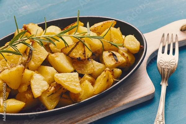 Fototapeta roasted potatoes iwith onion and rosemary in a cast iron pan over a white chopping board on a blue table
