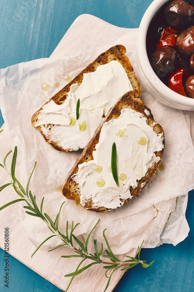 Fototapeta two slice of grilled bread with spreadable cheese and oil on a white chopping board, on a blue wooden table, with olives in a white bowl an rosemary