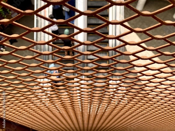 Fototapeta Metal mesh creates a grid - Close-up shot of a metal fence mesh grid with an escalator visible through it