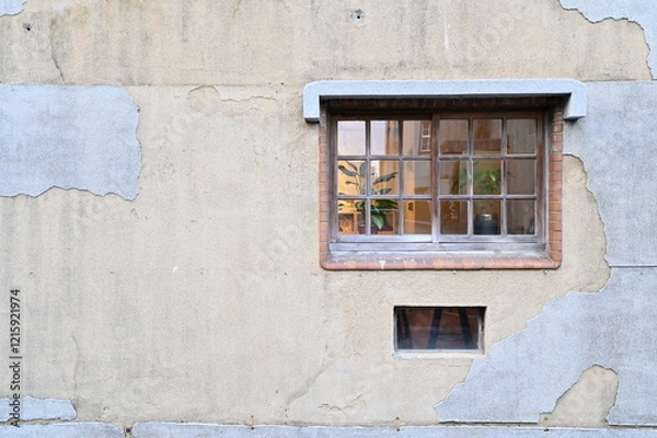 Fototapeta Against a timeworn wall with peeling paint, a red brick-framed glass window stands out. Inside, a soft yellow glow fills the room, while green plants near the window enhance the contrast of textures.