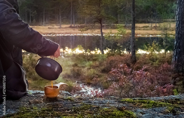 Fototapeta Pouring coffee from a kettle into a wooden cup near a lake in the forests