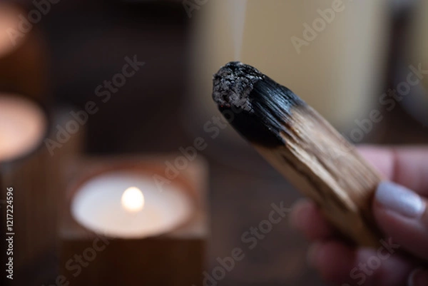 Fototapeta A stick of palo santo incense in a woman's hand smolders against the background of candles