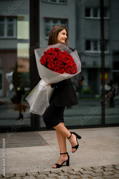 Fototapeta Gorgeous bouquet of red roses in hands of smiling girl in a black midi dress