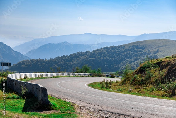 Obraz A mountain range with a winding road that is surrounded by trees. The road is curvy and has a white line painted on it. The mountains are covered in trees and the sky is blue