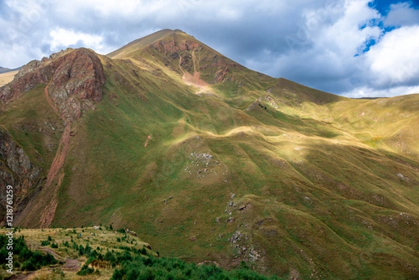 Obraz A mountain with a green hillside and a cloudy sky. The mountain is covered in grass and trees