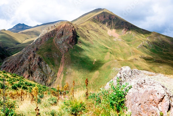 Obraz A mountain with a green hillside and a cloudy sky. The mountain is covered in grass and trees