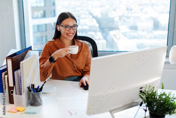 Fototapeta Good day at work. Happy European businesswoman drinking coffee at workplace, looking at computer monitor screen and smiling, free space