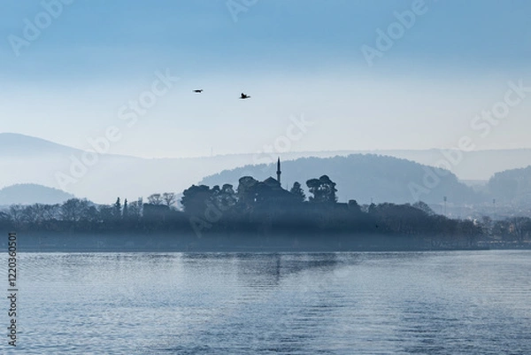 Fototapeta Early morning view of Ioannina lake. The minaret of Aslan Pasha Mosque and a cluster of trees rise above the mist, creating a striking contrast against the pale blue sky.
