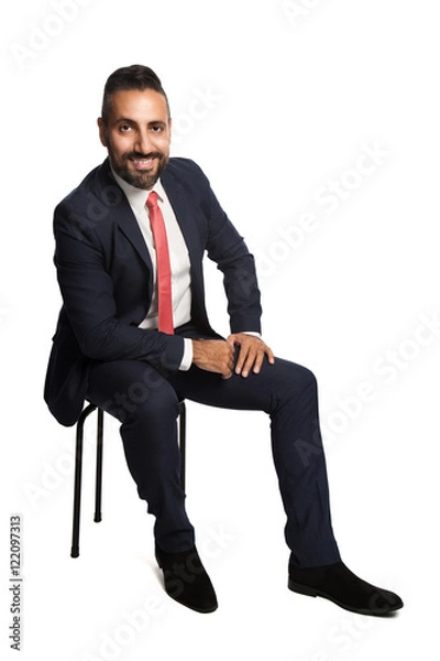 Fototapeta An attractive and smiling businessman wearing a blue suit and red tie, sitting down on a chair with hand on his knee. White background.
