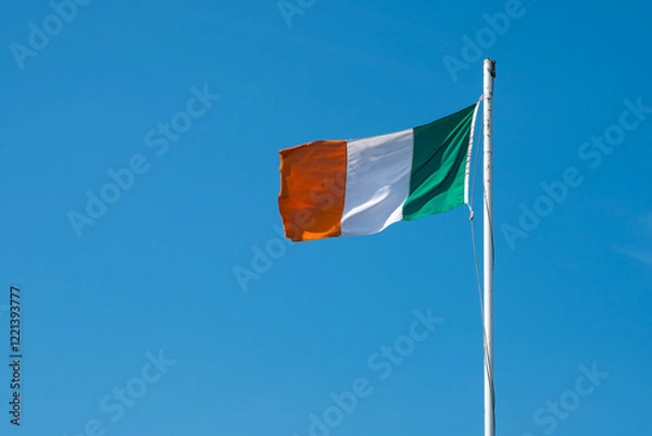 Fototapeta Irish flag waving in the wind on a flagpole under blue sky background. Ireland national flag symbol flying on a pole