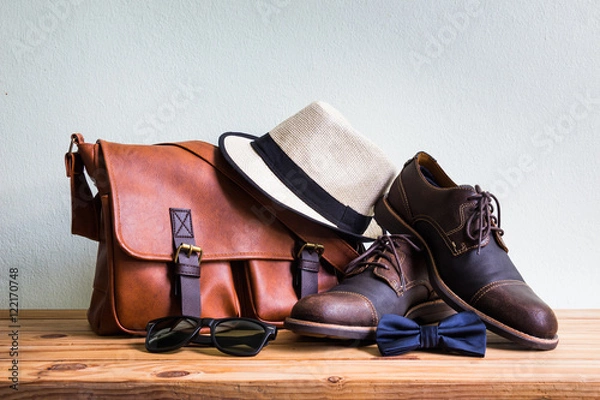 Fototapeta Akcesoria męskie z brązową skórzaną torbą, brązowymi butami, klasycznym kapeluszem, okularami przeciwsłonecznymi i niebieską muszką na drewnianym stole na tle ściany