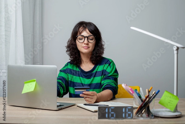 Fototapeta A woman wearing glasses and a green-striped shirt sits at a desk with a laptop. She holds a credit card in her hand and smiles while looking at the screen. The workspace is neat and modern