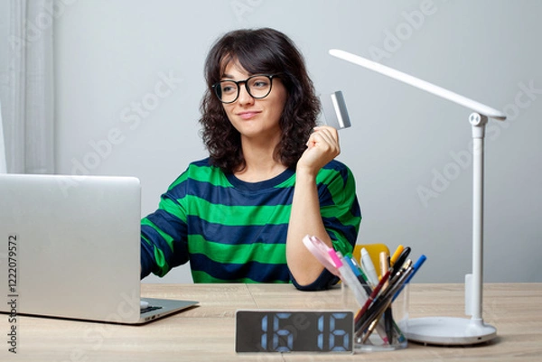 Fototapeta A woman wearing glasses and a green-striped shirt sits at a desk with a laptop. She holds a credit card in her hand and smiles while looking at the screen. The workspace is neat and modern