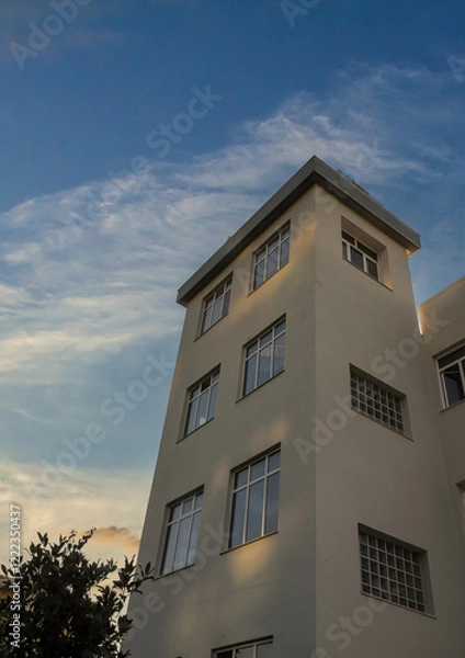 Fototapeta Tall building with modern architecture stands against a clear sky during sunset, showcasing shadows and reflections from nearby foliage