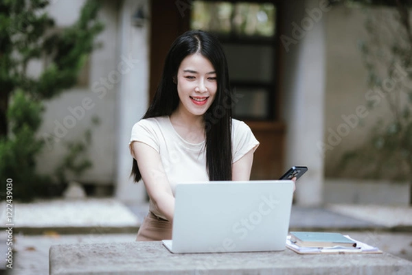 Fototapeta Cheerful Asian woman working in a coffee shop using her laptop. She enjoys teleworking, studying or surfing the internet to relax.