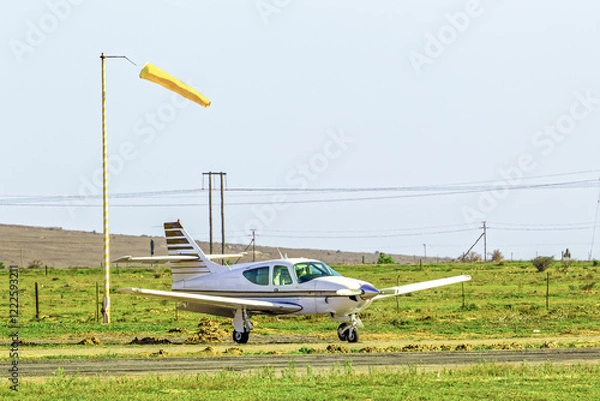 Obraz White and brown four-seat single engine light aircraft parked next to windsock at a small airstrip in the Western Cape, South Africa