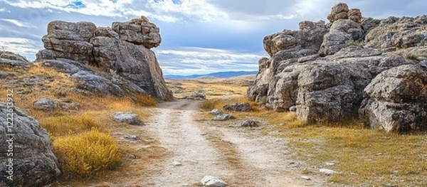 Fototapeta Dirt path winding through rocky cliffs with gray boulders and golden grass under a cloudy sky creates an adventurous exploration scene.