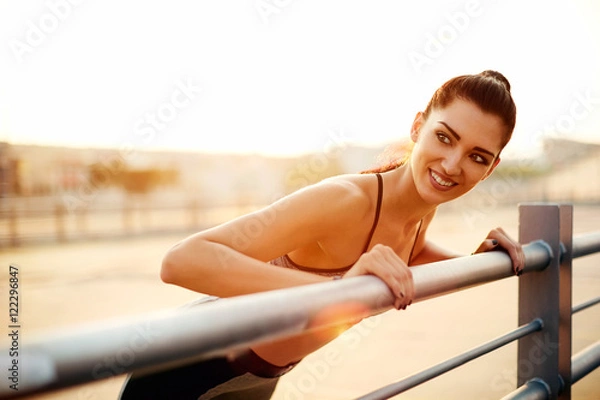 Fototapeta Portrait of a young smiling female runner leaning on the railing satisfied with workout