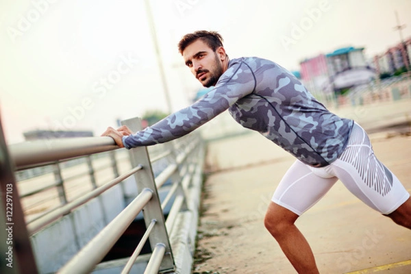Obraz Male runner doing stretching exercise, preparing for sunset workout