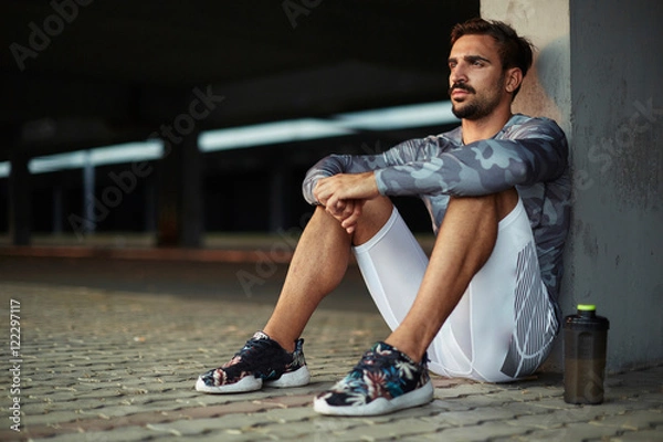Fototapeta Portrait of a tired but satisfied with his training men runner resting after workout while sitting on concrete