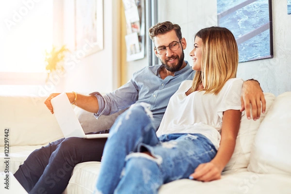 Fototapeta Young Couple Sitting On Sofa Using Laptop looking at each other and smiling