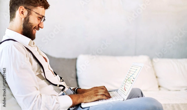 Fototapeta Happy young man, wearing glasses and smiling, as he works on his laptop to get all his business done early in the morning