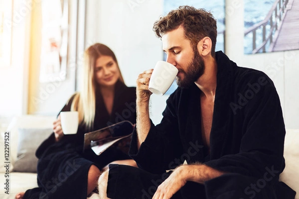 Fototapeta Happy young couple sitting on sofa in the bathrobe and drinking coffe in the morning