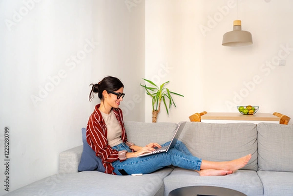 Fototapeta Peaceful home setting. A young woman smiling while working on her laptop in a well-lit living room. Minimalist and lifestyle concept
