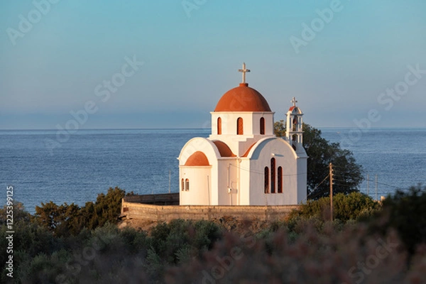 Fototapeta A typical Cretan chapel by the coast, with white walls, a red-tiled roof, and a backdrop of blue sea and sky.