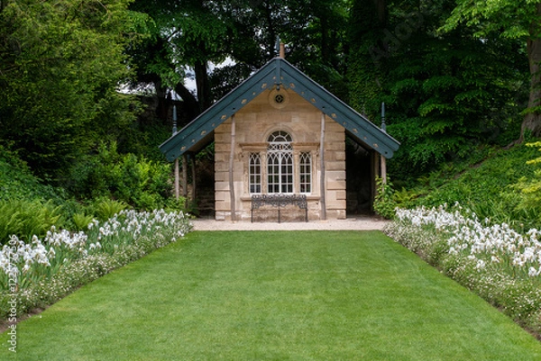 Fototapeta A small house with a green roof sits in a lush green field. The house is surrounded by a beautiful garden with white flowers. The garden is well-maintained and adds to the overall charm of the scene