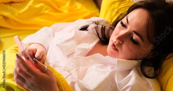 Fototapeta A young Caucasian woman in a white T-shirt uses a smartphone while lying on a bed with yellow bedsheets. Suitable for lifestyle, technology, and relaxation-themed videos.