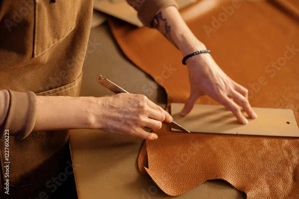 Obraz Close-up of hands skillfully cutting leather with a blade, showing detailed craftsmanship. Table covered with pieces of leather and tools for crafting