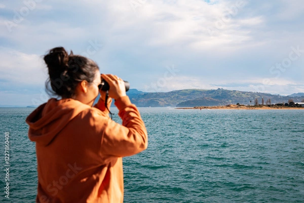 Obraz Woman in an Orange Hoodie Using Binoculars to Observe Coastal Landscape in Santoña, Cantabria, Spain During Springtime
