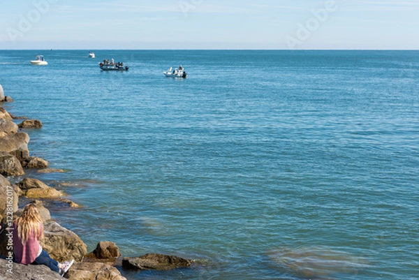 Obraz A woman with long blond hair is seen from behind sitting on a rock jetty looking out to sea where there  people fishing on boats,  Ponce Inlet, Florida, USA