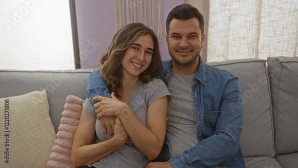 Fototapeta Man and woman couple sitting together on a couch in a cozy living room indoors, smiling and embracing each other warmly.