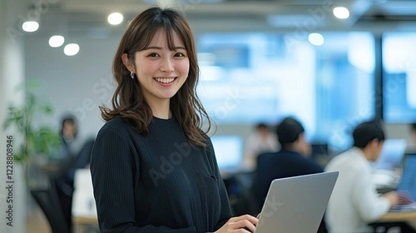Obraz Smiling Japanese businesswoman standing in an office, holding her laptop. She is wearing casual clothes, and other people working behind her are blurred.,generated ai. 

