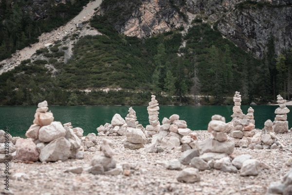 Fototapeta A serene landscape featuring multiple balanced stone cairns on a rocky shore, overlooking a beautiful turquoise lake surrounded by lush mountains. The image captures a sense of harmony, nature.