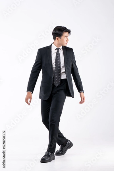 Fototapeta A man in a black suit and tie stands in front of a white background