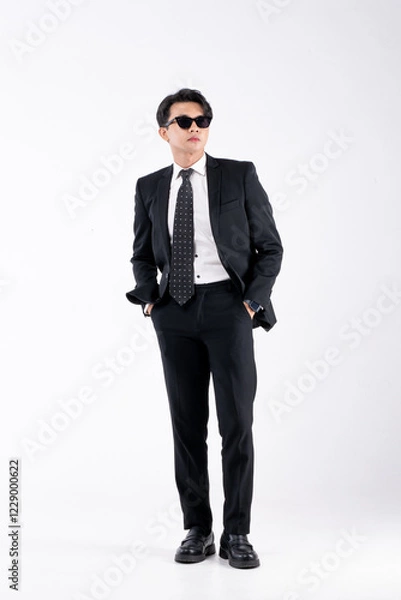 Fototapeta A man in a black suit and tie stands in front of a white background