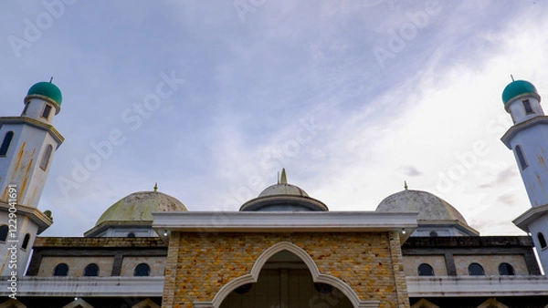 Fototapeta A low-angle, eye-level shot that captures the grandeur of the mosque's exterior against a partly cloudy, clear sky. The mosque showcases its architecture with its two towering minarets on either side.