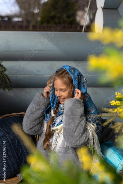 Fototapeta A beautiful Russian girl in a fur coat and headscarf sits on the porch of a wooden house on a sunny spring day.Family values,traditions and home comfort.The girl covers her hair with a beautiful scarf