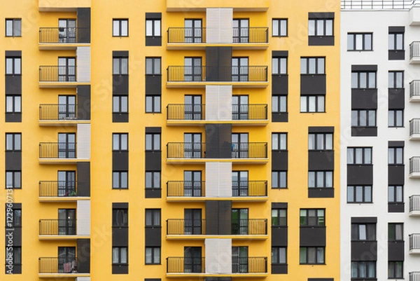Fototapeta Modern apartment buildings with yellow and white facades, large windows, and balconies with metal railings, against a cloudy sky, front view