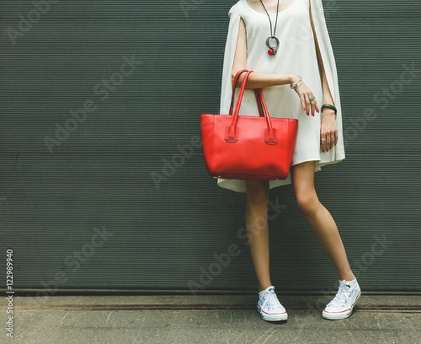 Obraz Fashionable beautiful big red handbag on the arm of the girl in a fashionable white dress, posing near the wall on a warm summer night. Warm color.