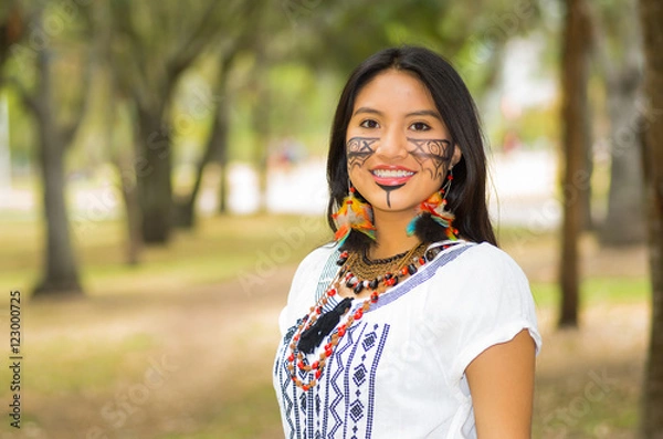 Obraz Beautiful Amazonian woman with indigenous facial paint and white traditional dress posing happily for camera in park environment, forest background