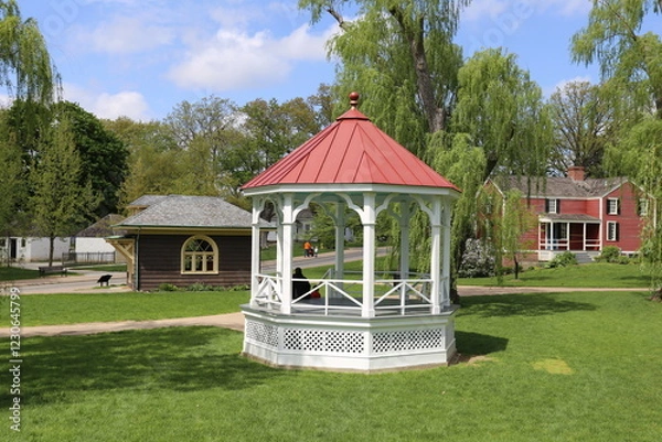 Fototapeta White Gazebo with Red Roof in a Green Park Surrounded by Trees and Historic Homes